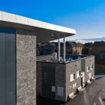 Aerial view of the rear of a recreational building with outdoor picnic tables, showing the gray ACM and stone exterior.