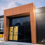 The front entrance of a modern commercial building featuring a black and metallic brown ACM facade under a bright sky.