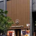 Paris Baguette store front facade with brown aluminum panels and green foliage foreground.