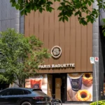 Paris Baguette store front with brown aluminum panels, a parked black SUV, and foliage.