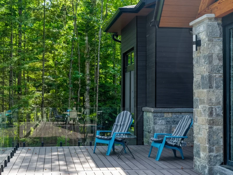 Outdoor patio with composite decking, glass railing, blue Adirondack chairs, and a house wall finished with anthracite cladding and stone.
