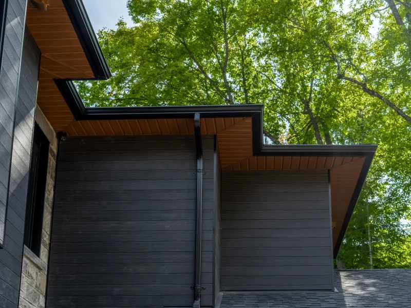 Close-up of modern roofline showing the dark anthracite cladding wall, black gutter, and contrasting orange-brown wood soffit.