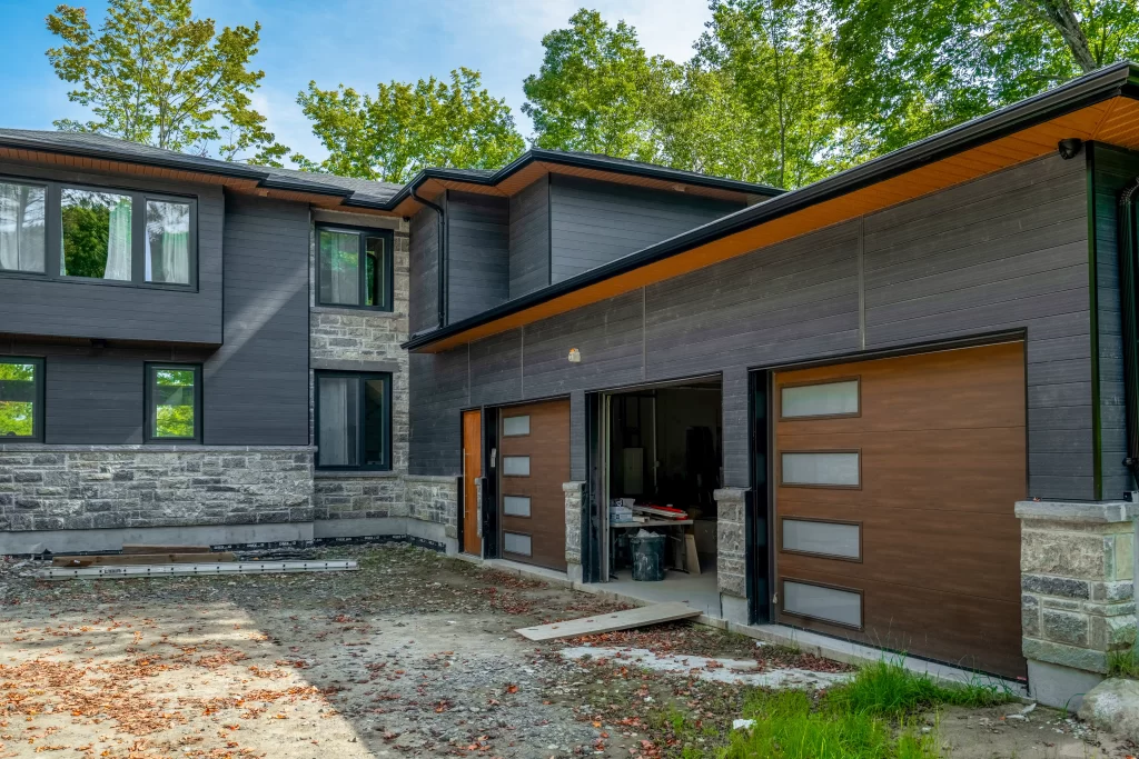 Expansive modern home with two garage bays featuring faux wood doors, stone veneer base, and dark anthracite cladding on the upper levels.