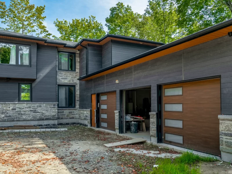 Expansive modern home with two garage bays featuring faux wood doors, stone veneer base, and dark anthracite cladding on the upper levels.
