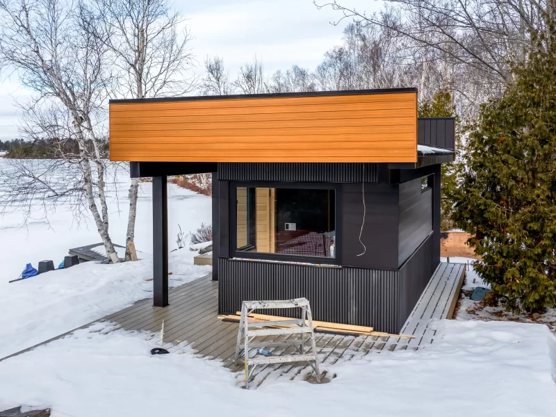 Small lakefront building or sauna with dark anthracite composite cladding and a prominent horizontal band of wood-look cladding in the snow.