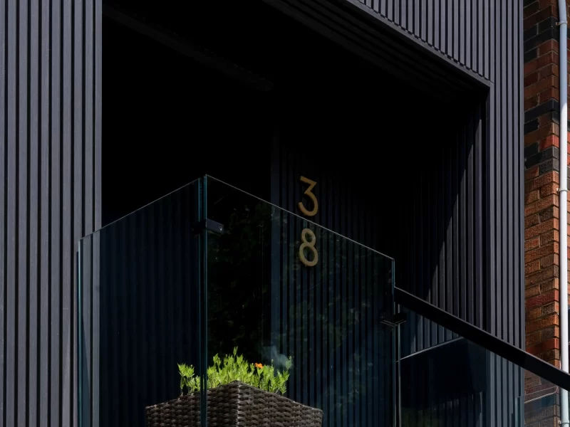 Close-up of a modern front porch entry framed by vertical anthracite siding, featuring a glass railing, potted plant, and house number 38.