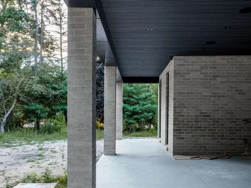 Covered outdoor walkway featuring heavy gray brick columns and a linear anthracite soffit with a black fascia, next to a wooded area.