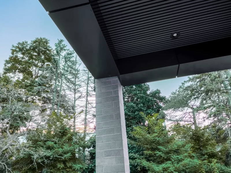 Close-up of a modern gray brick column meeting a black anthracite soffit and fascia, with tall trees and blue sky visible.