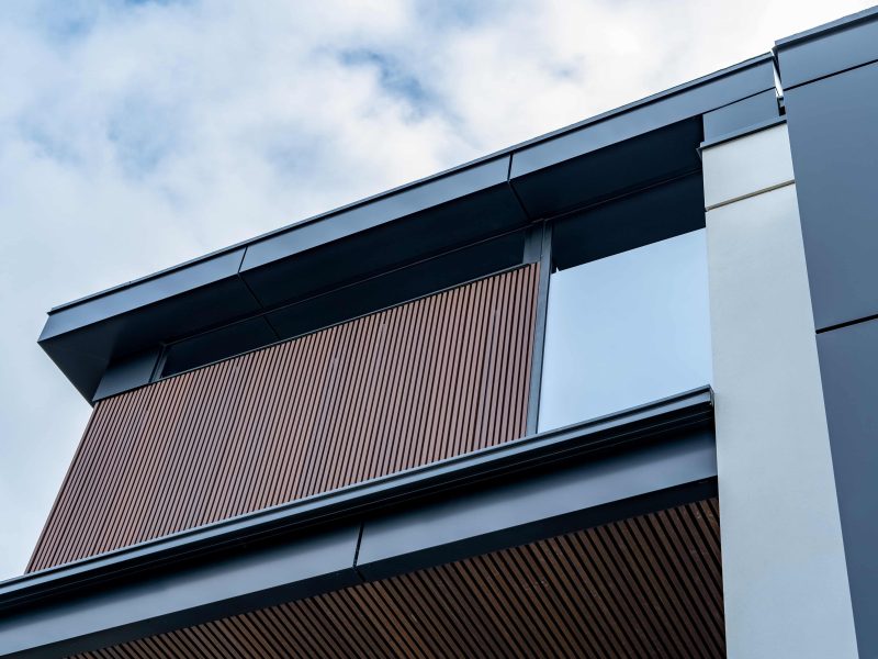 Close-up of brown vertical fluted siding under a black metal soffit against a bright, cloudy sky.