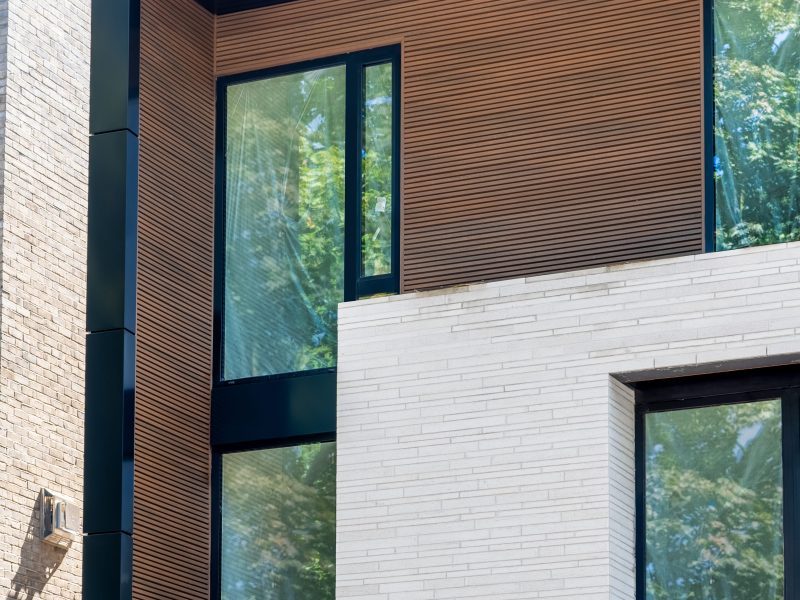 Modern facade section with brown fluted siding, white brick, and black metal trim around large windows.